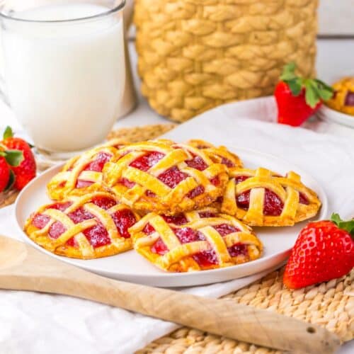 A plate of small lattice-topped strawberry pies sits on a woven mat with fresh strawberries, a wooden spoon, and a glass of milk nearby.