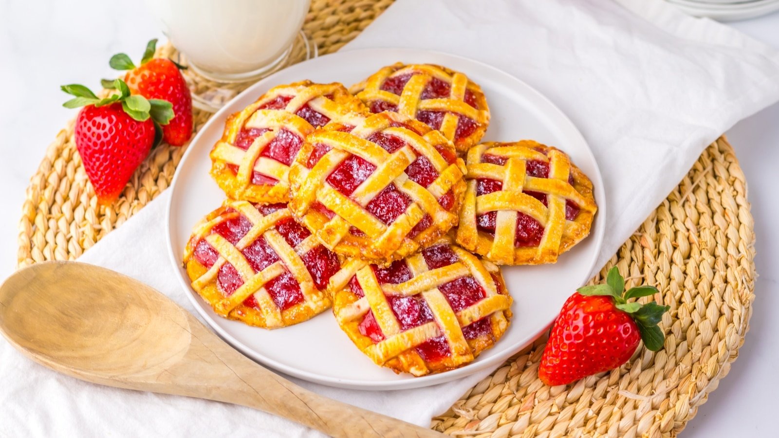 A plate of lattice-topped strawberry pastries on a woven mat with fresh strawberries and a wooden spoon nearby. A glass of milk is in the background.