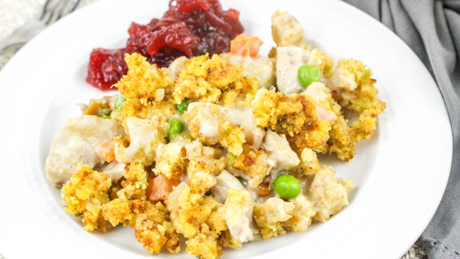 A plate of turkey casserole with peas and carrots, topped with breadcrumbs, served with cranberry sauce on the side.