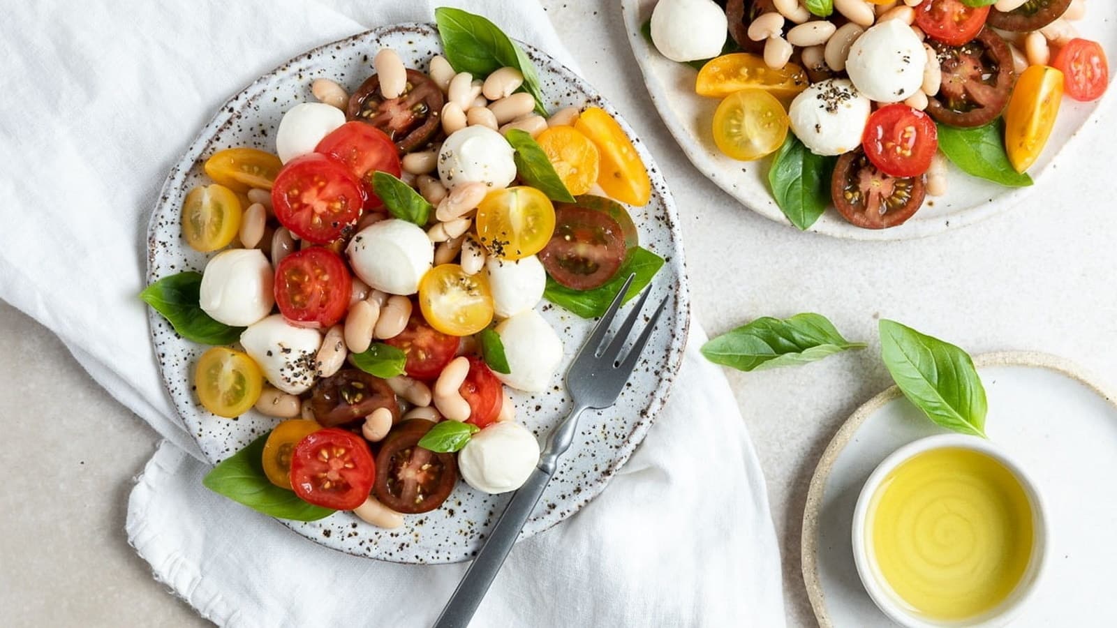 Two plates containing a salad with cherry tomatoes, mozzarella, white beans, and basil. A fork rests on one plate. A small dish of olive oil is nearby on a white cloth.