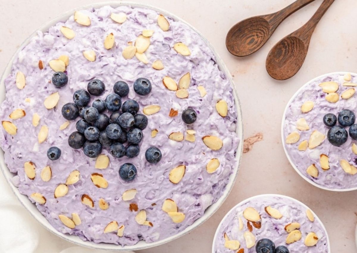 A bowl of purple fruit salad topped with blueberries and sliced almonds hints at summer freshness—perfect inspiration for your next brunch or to add to your list of 12 Tasty Blueberry Recipes. Two smaller bowls and wooden spoons sit nearby.