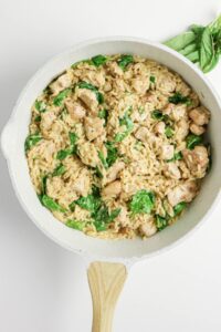 A white skillet filled with Lemon Chicken Orzo Pasta, featuring tender orzo, spinach, and seasoned chicken, garnished with fresh basil leaves, on a white background.
