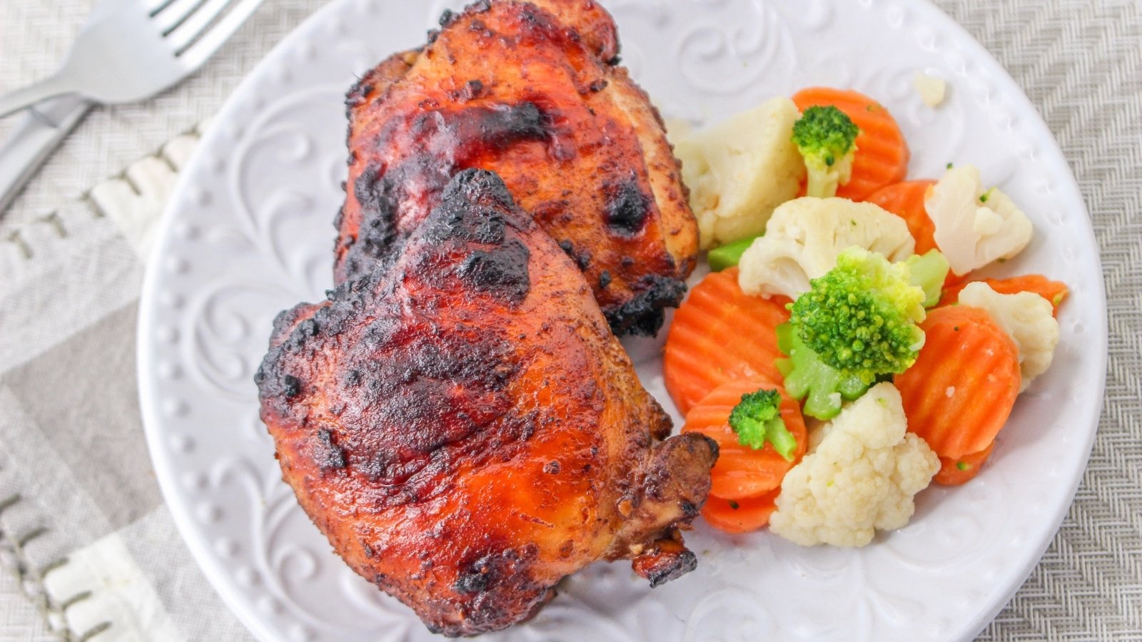 Chicken thighs and steamed vegetables on a white plate, placed on a gray tablecloth with a fork and napkin nearby.