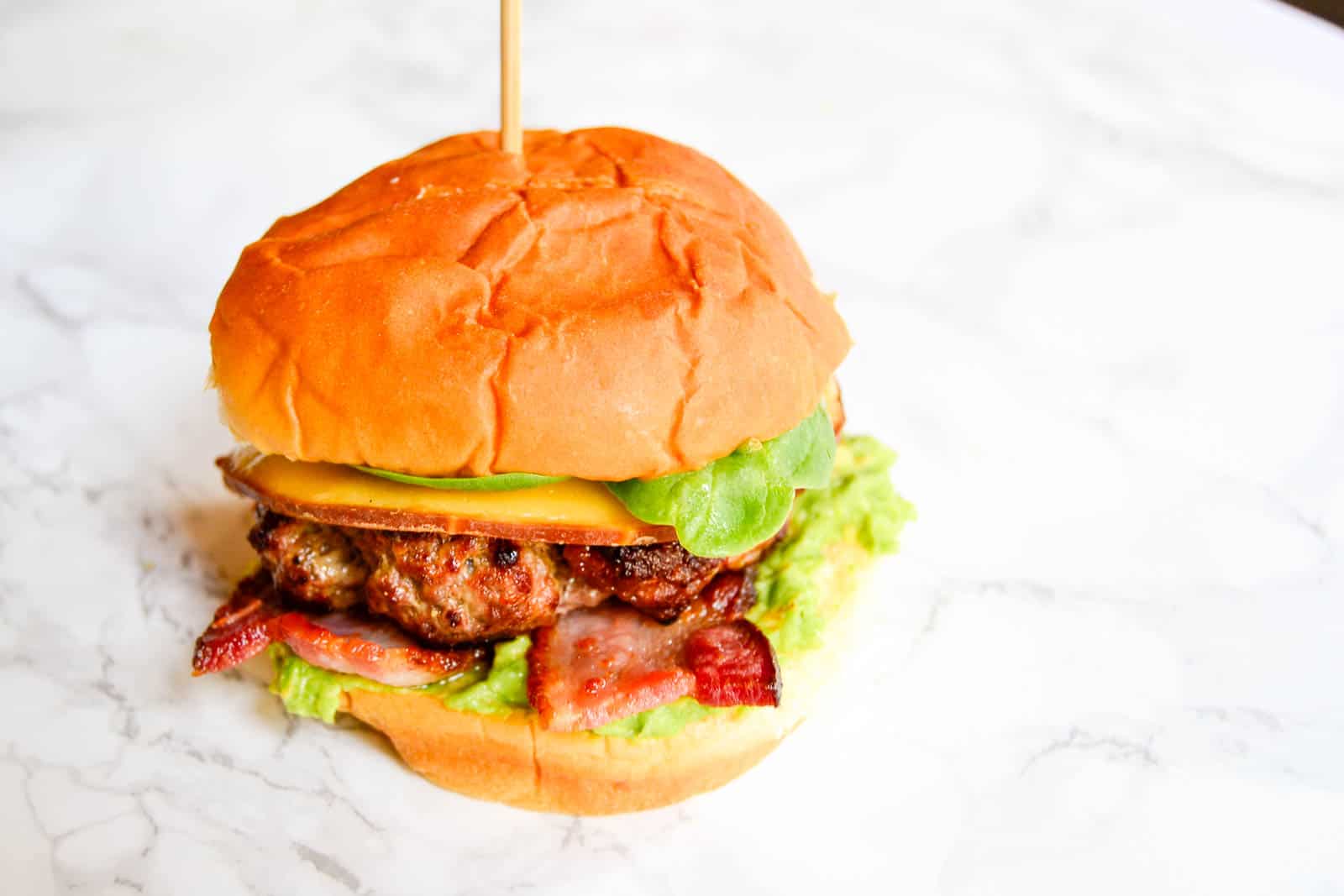 A burger with a beef patty, cheese, bacon, lettuce, and a slice of avocado on a bun, held together by a skewer, placed on a marble surface.