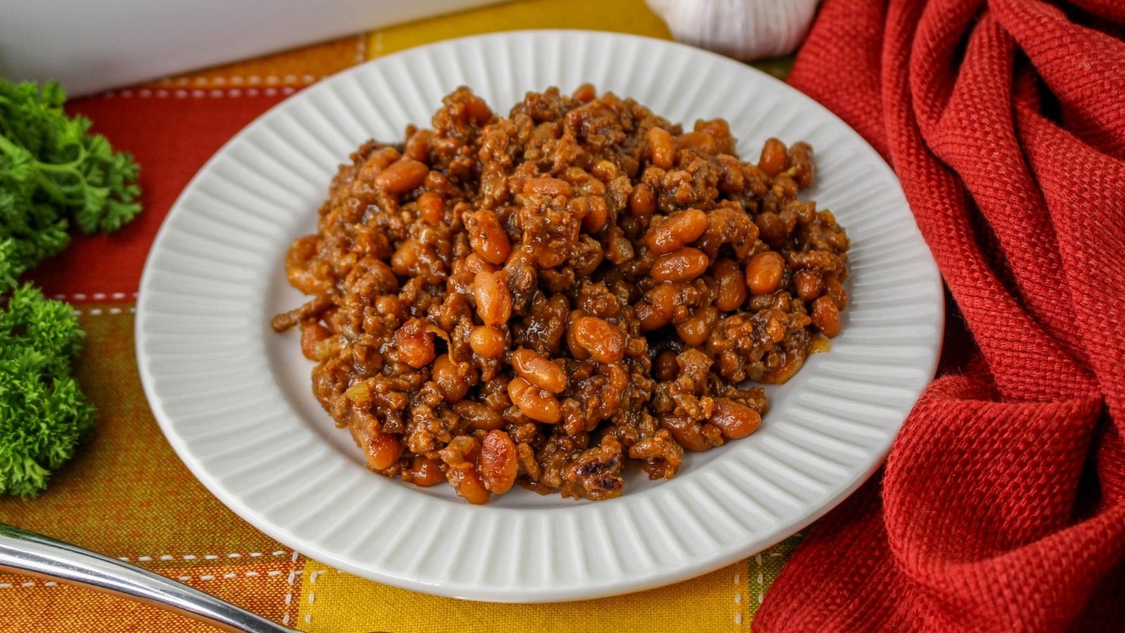A plate of baked beans mixed with ground beef, placed on a white plate. The plate is on a colorful tablecloth, with a red cloth and greens nearby.