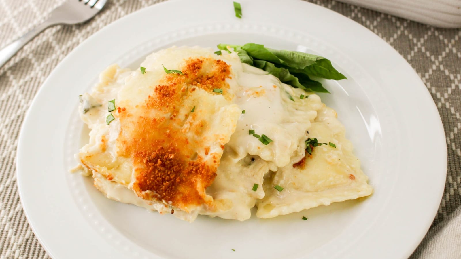 Plate of creamy ravioli topped with a sprinkling of browned cheese, garnished with basil, on a white dish with a fork nearby.
