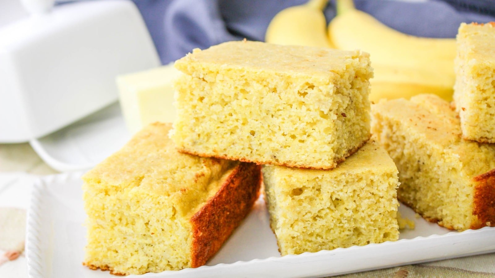 Slices of cornbread stacked on a white plate with bananas in the background.