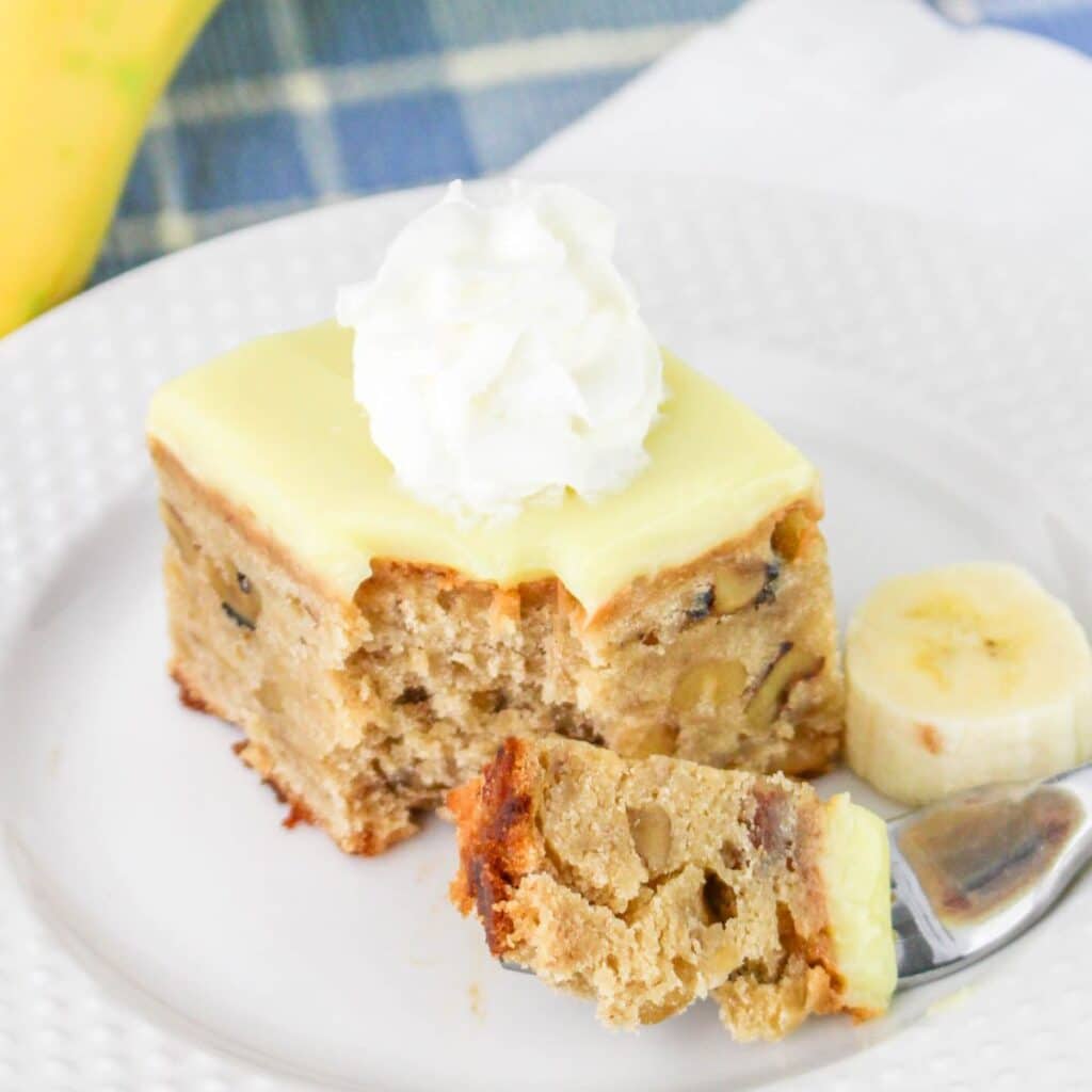 Slice of banana cake with frosting and whipped cream on a white plate, accompanied by banana slices.