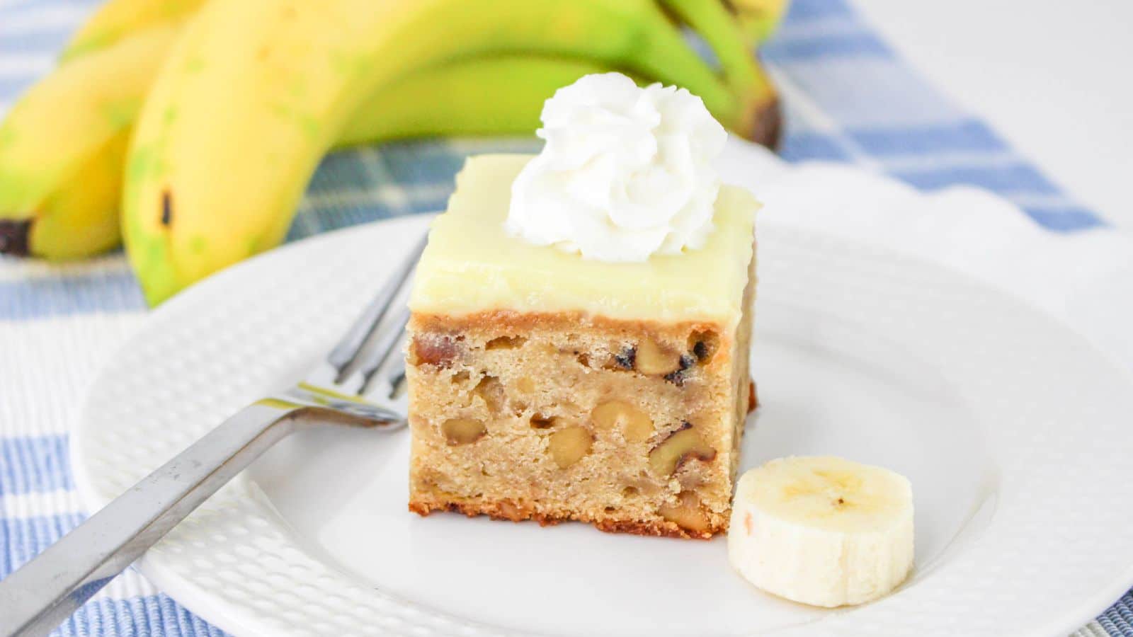 A slice of banana cake with nuts, topped with frosting and whipped cream, on a white plate. A banana slice and fork are beside it. Whole bananas are in the background.