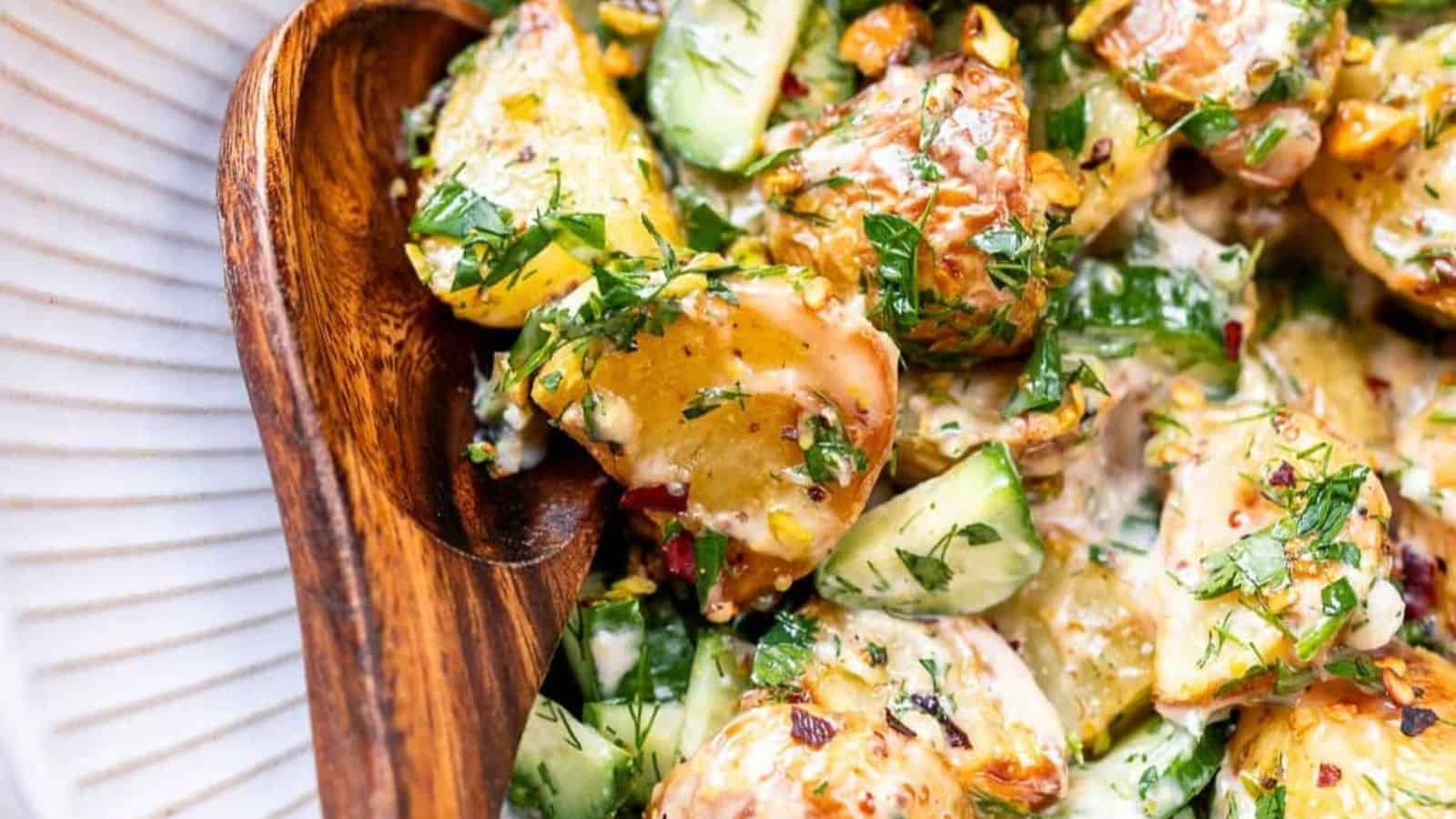 Wooden bowl with a creamy potato salad, featuring chopped herbs, cucumbers, and corn on a white plate.