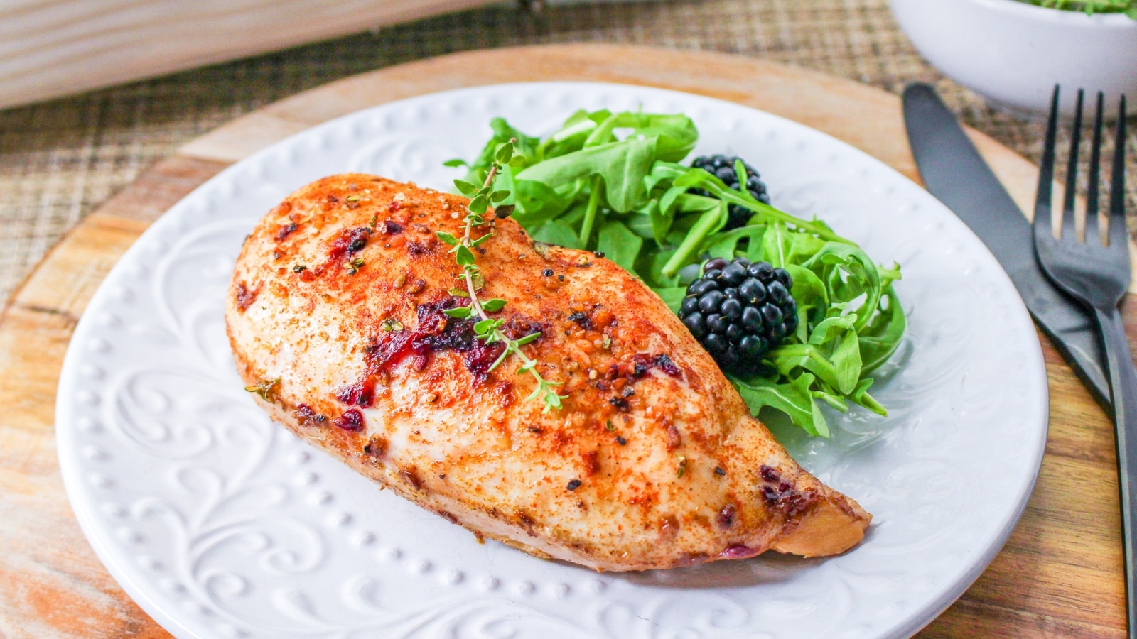 Grilled chicken breast garnished with herbs, served with leafy greens and blackberries on a white plate. A knife and fork are placed beside it.