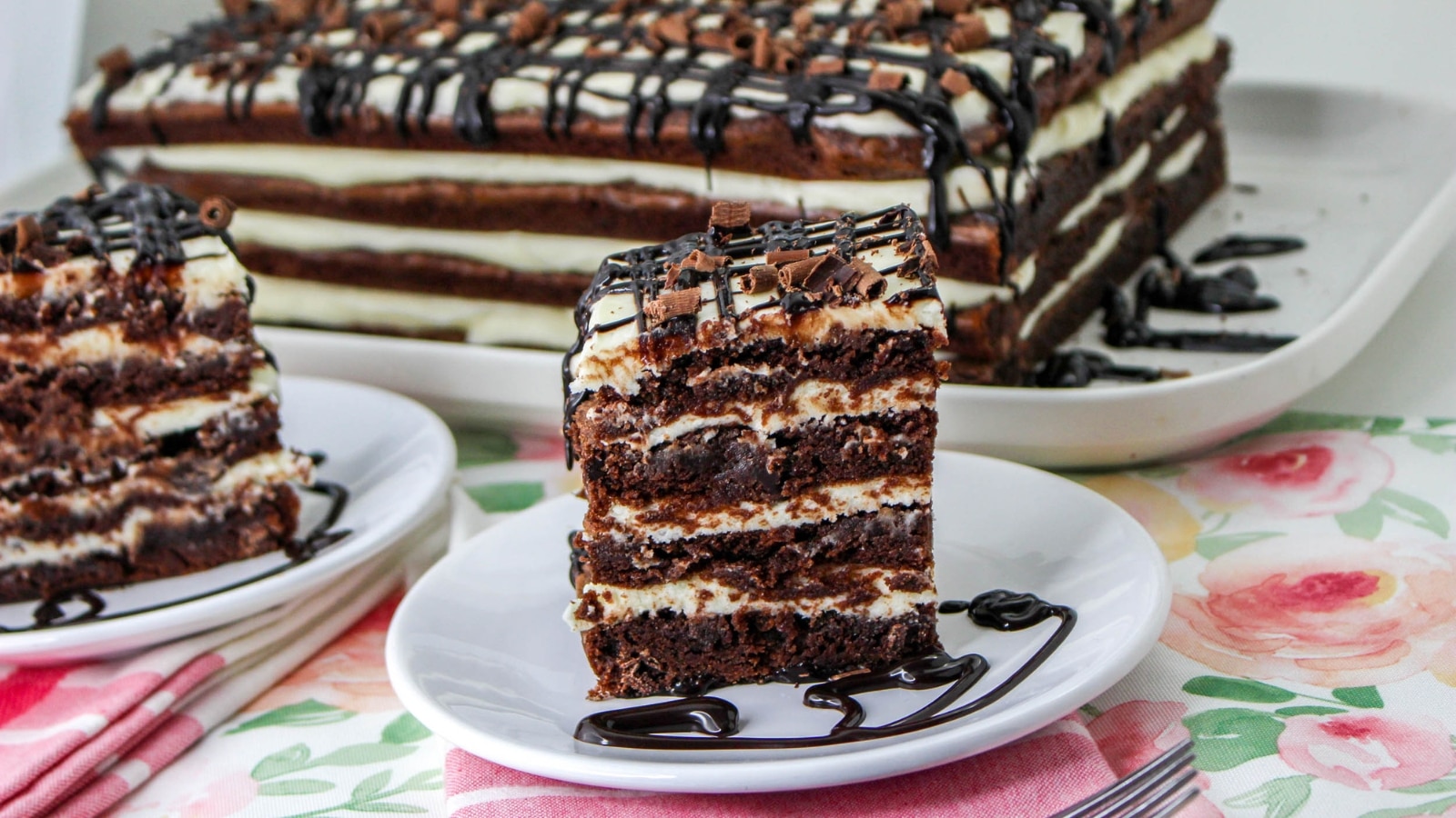Slice of layered chocolate and cream cake on a white plate with chocolate drizzle, resting on a floral tablecloth. Another cake slice and entire cake are in the background.