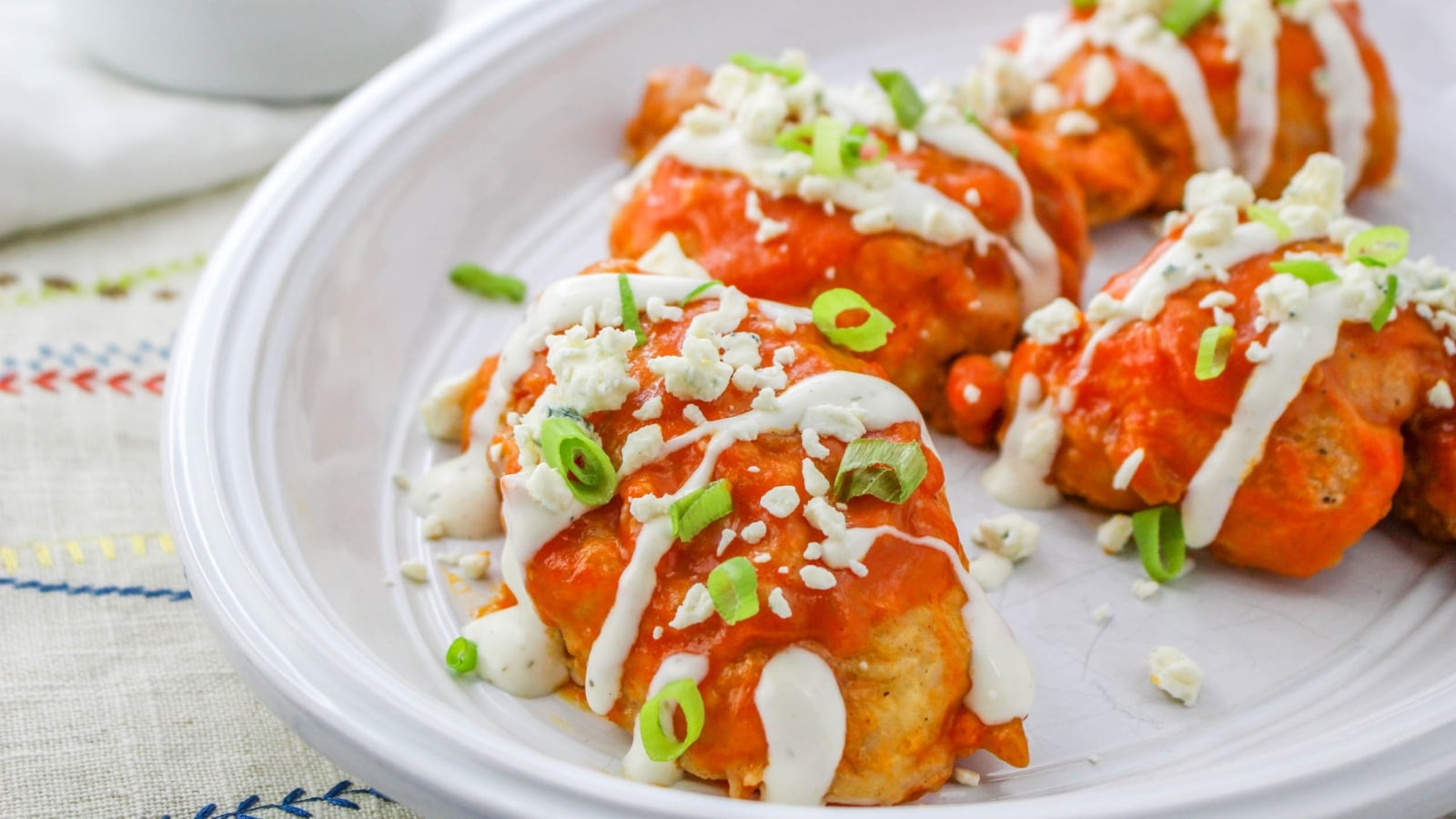 Buffalo chicken bites drizzled with white dressing, topped with chopped green onions and crumbled cheese, served on a white plate.