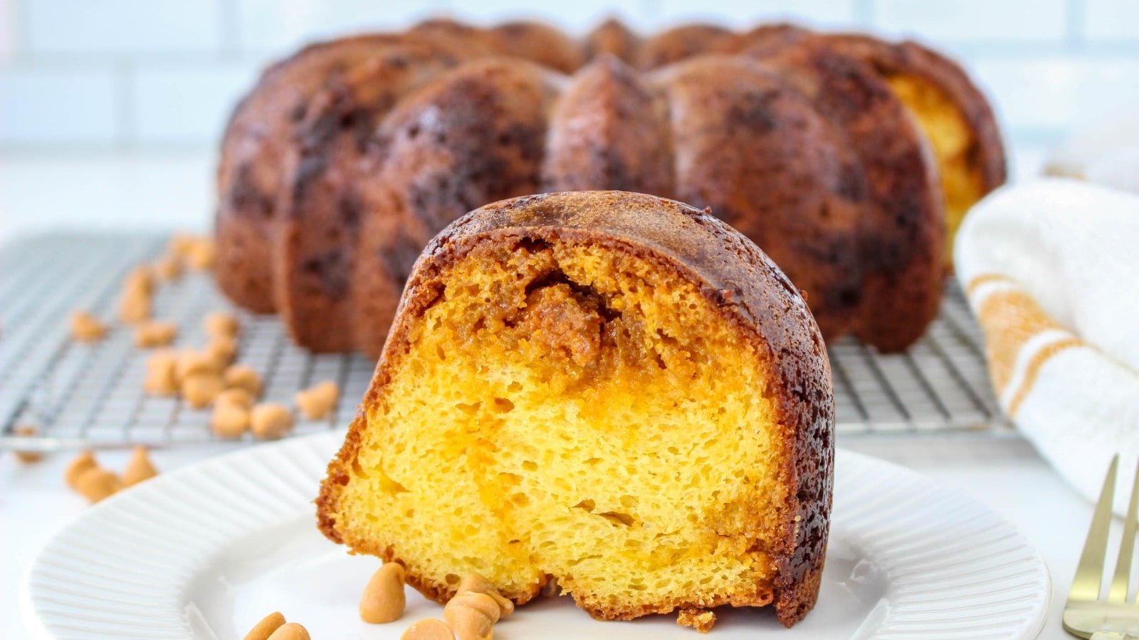A slice of bundt cake on a plate with caramel chips, with the whole cake in the background on a cooling rack.