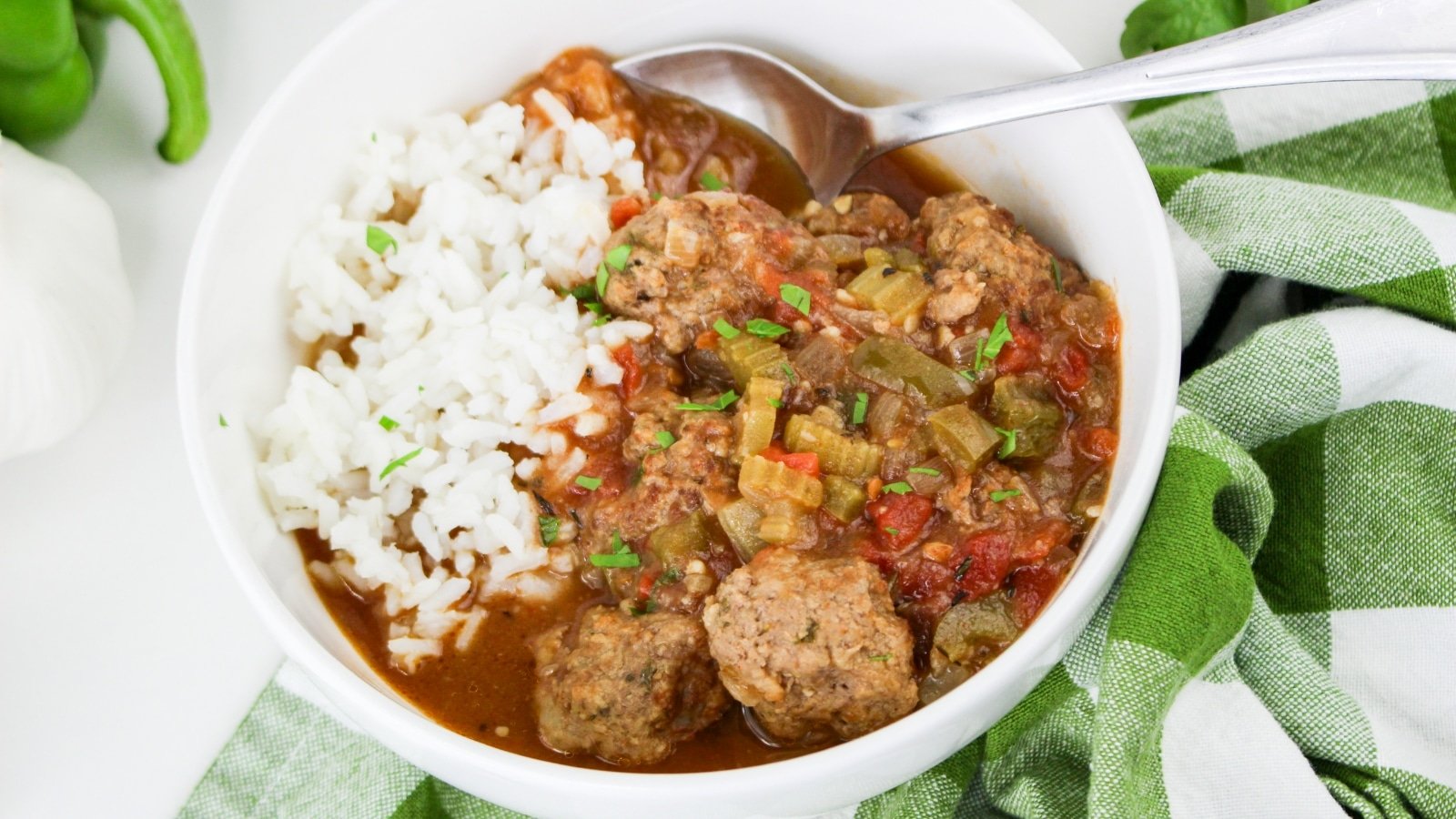 A bowl of rice and meatball stew with vegetables, served with a spoon, placed on a green checkered cloth.