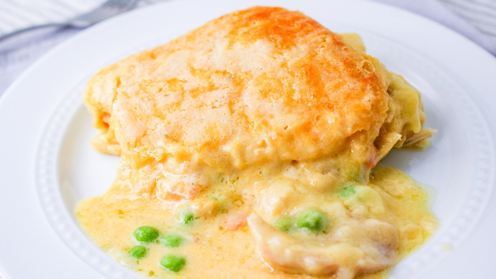 A serving of chicken pot pie with a golden-brown crust on a white plate. Visible peas and creamy filling are partially oozing out.