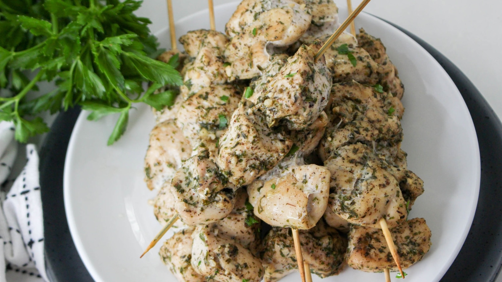Grilled chicken skewers seasoned with herbs on a white plate, accompanied by a bunch of fresh parsley on the side.