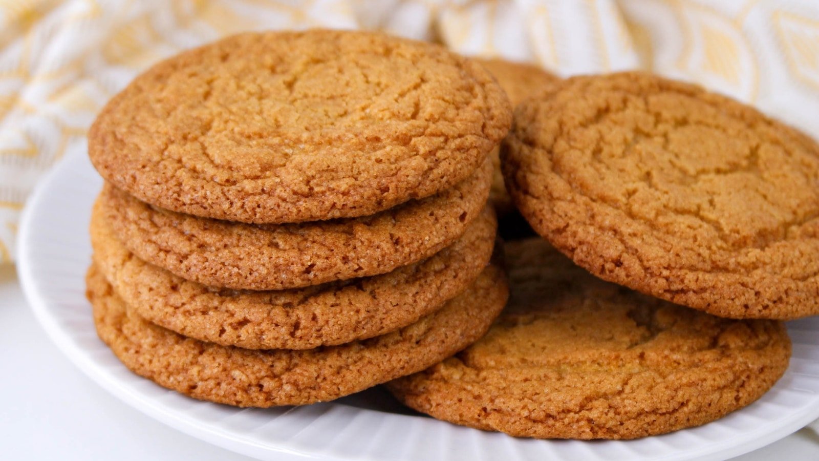 A stack of golden-brown cookies on a white plate with a yellow and white cloth in the background.