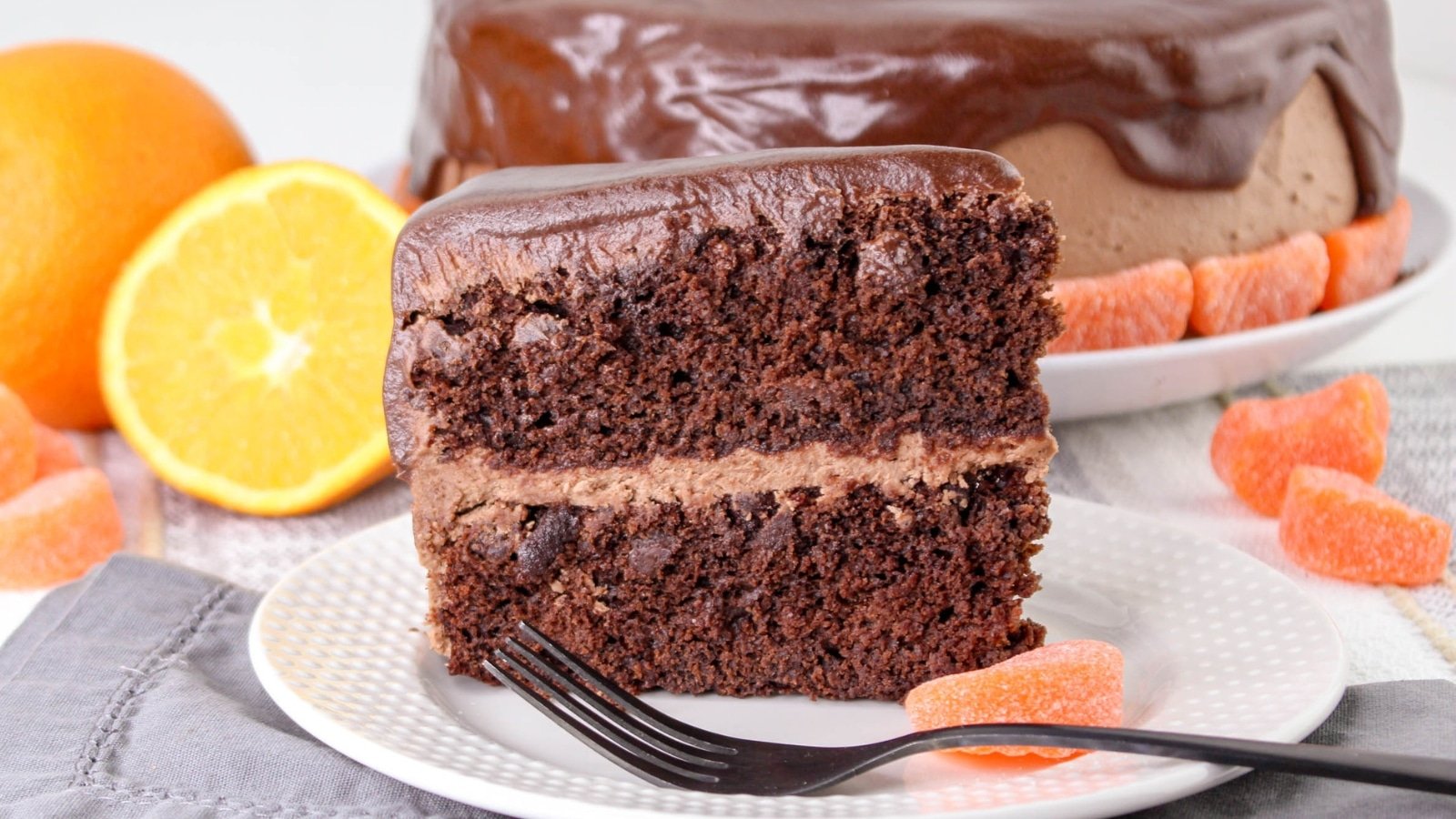 Slice of chocolate cake with chocolate frosting on a white plate, garnished with orange slice and candy. A whole cake is in the background.