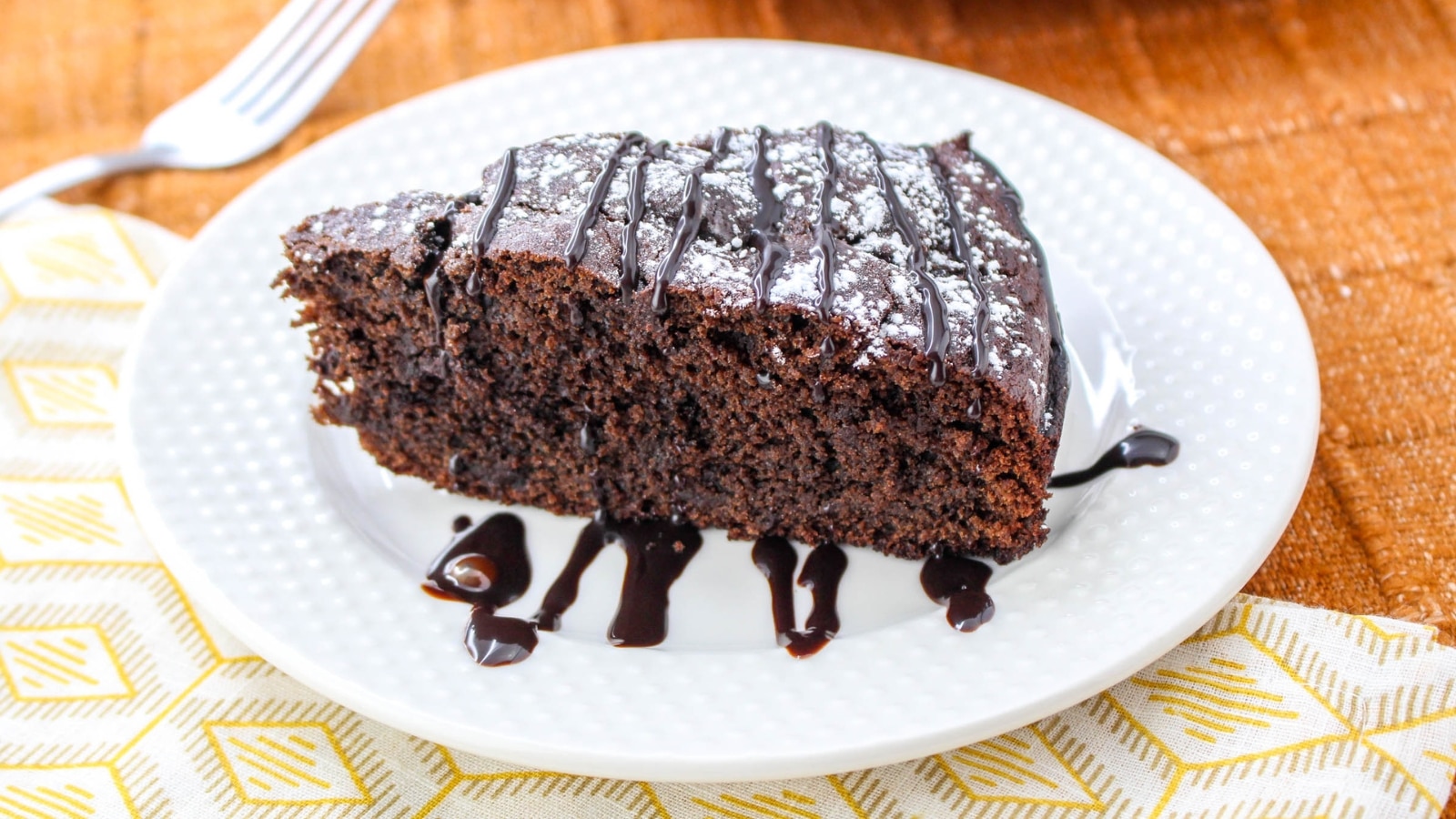 A slice of chocolate cake with chocolate drizzle and powdered sugar on a white plate, placed on a patterned napkin, with a fork nearby.
