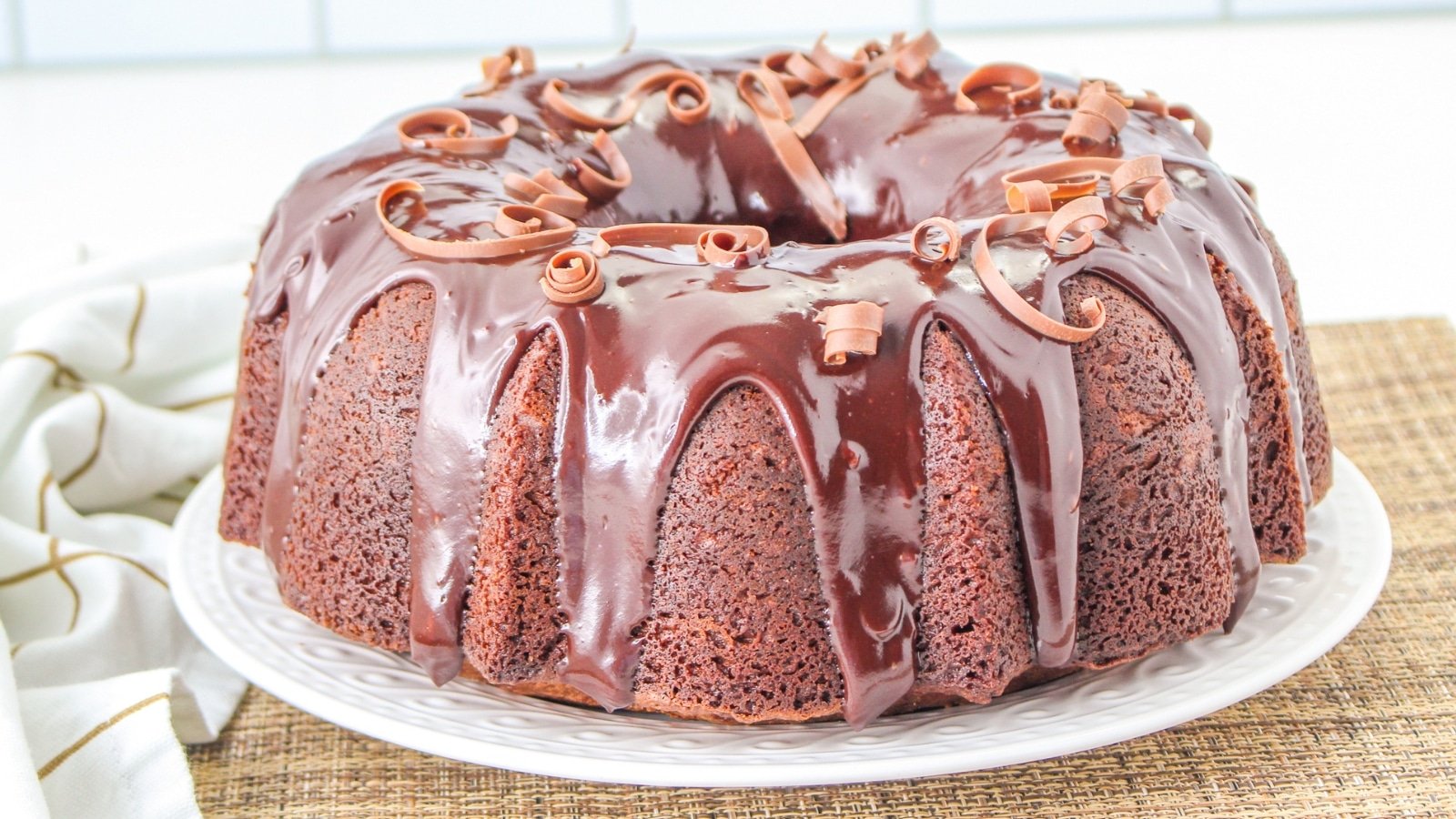 Chocolate bundt cake with glossy chocolate glaze and decorative chocolate curls on a white plate.