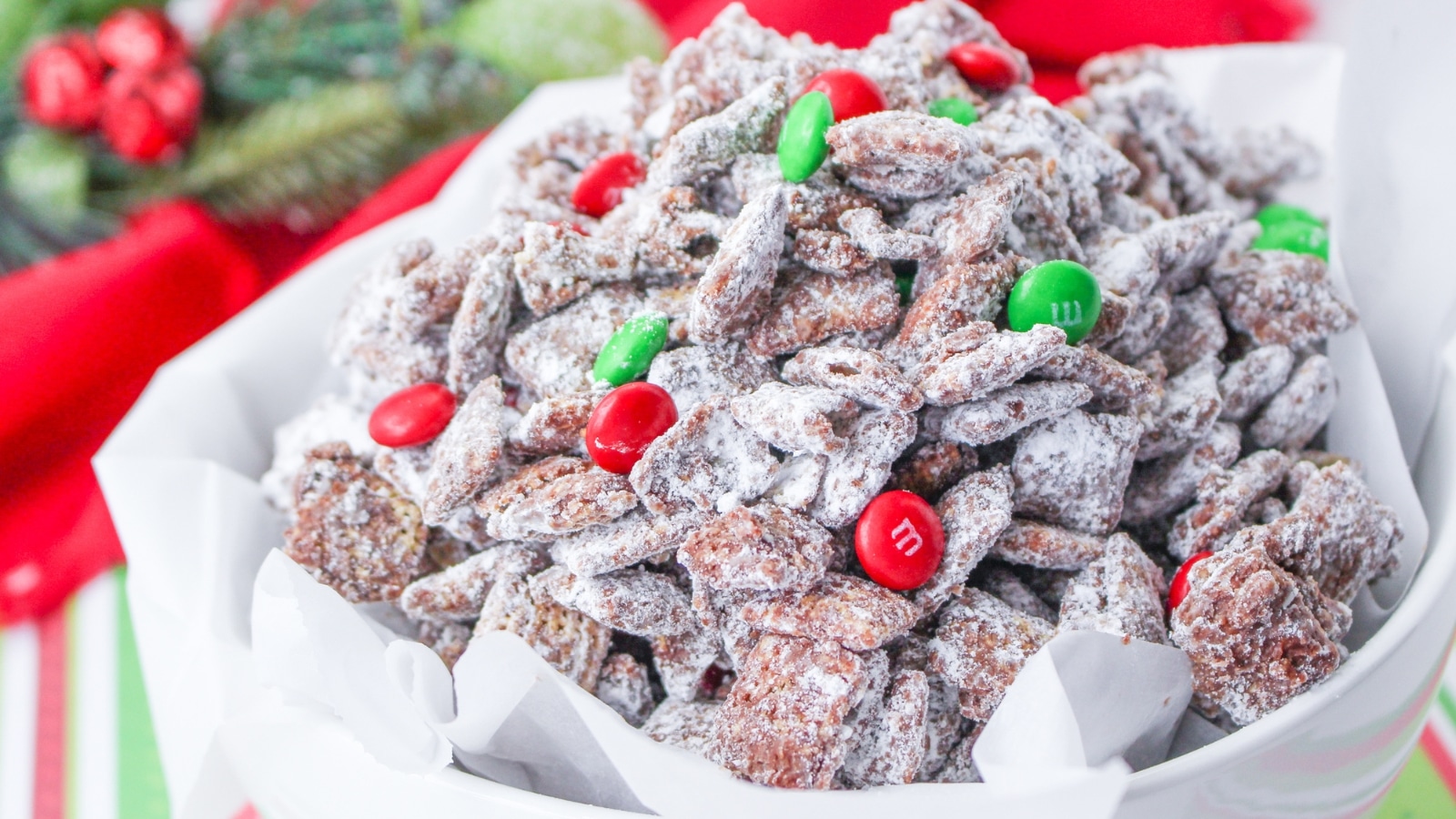 Bowl of coated snack mix with powdered sugar and red and green chocolate candies on a festive decorated table.
