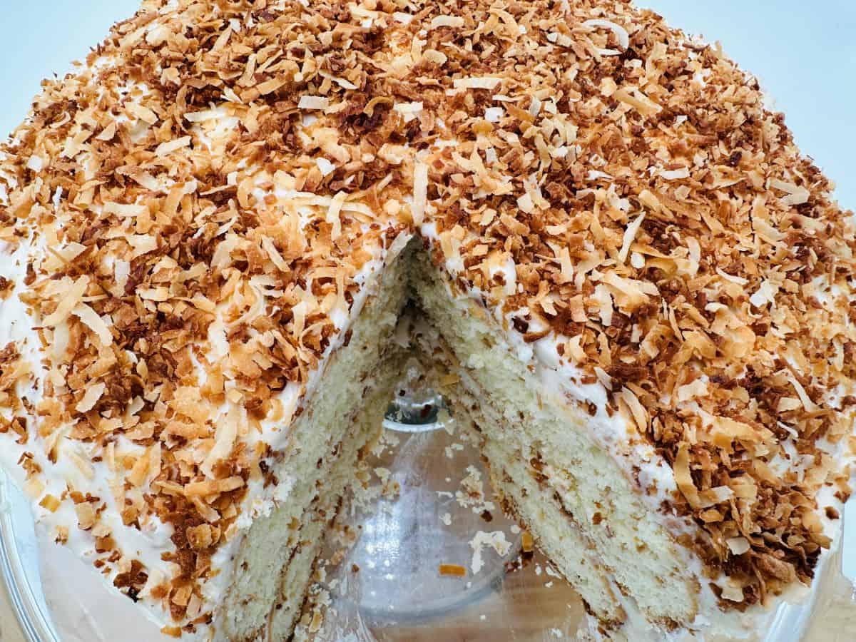 Round cake with white frosting and toasted coconut flakes on top, one slice missing, displayed on a glass cake stand.
