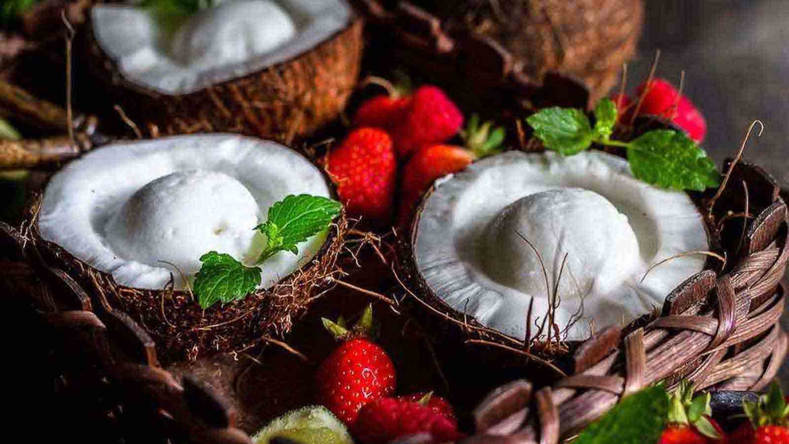 Halved coconuts filled with white dessert, garnished with mint leaves, surrounded by strawberries and small citrus fruits in a woven basket.
