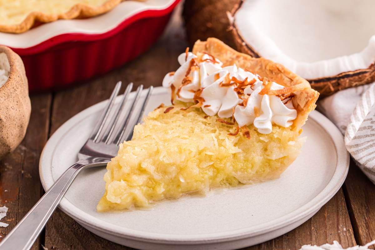 A slice of coconut cream pie topped with whipped cream and toasted coconut sits on a plate with a fork; just one of 15 coconut creations, with a whole pie and a coconut tempting in the background.