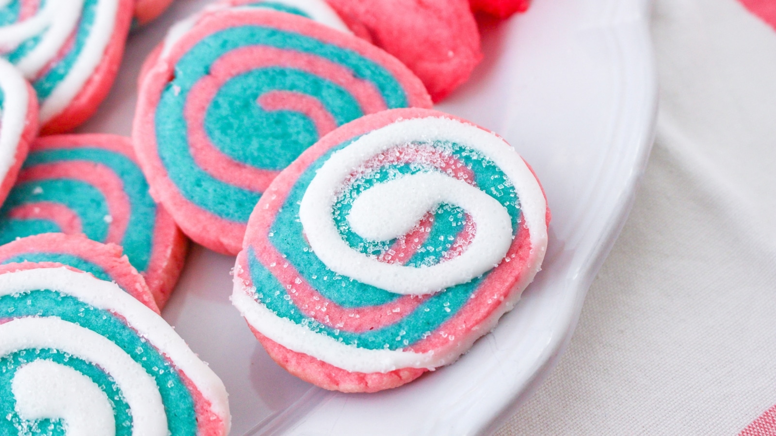 Close-up of spiral cookies with pink, blue, and white swirls on a white plate. The cookies are covered with a light sprinkle of sugar.
