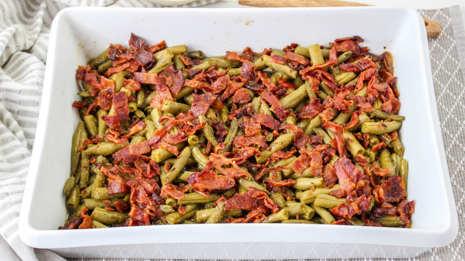 A white baking dish filled with cooked green beans mixed with crispy bacon pieces.