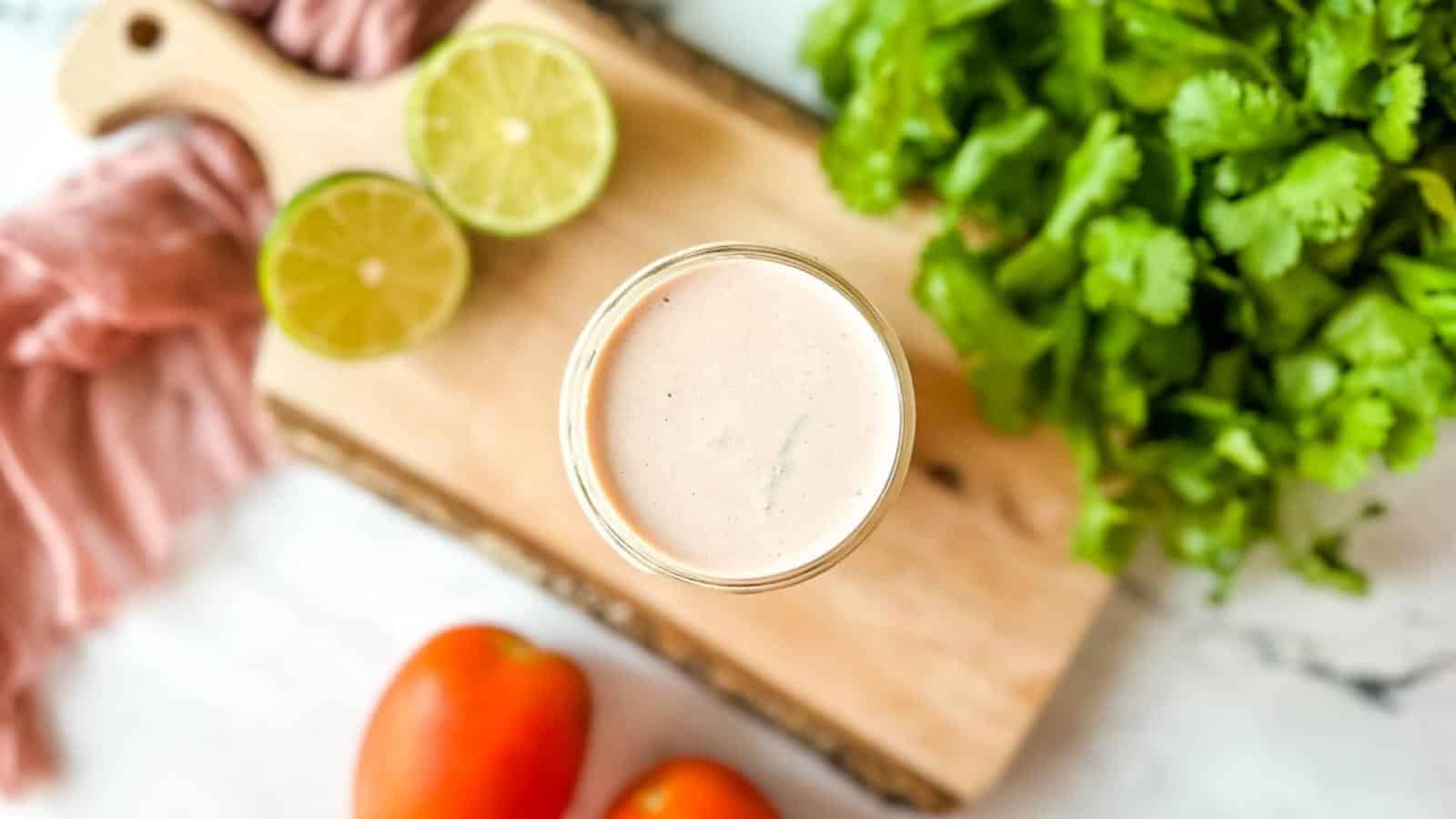 A jar of creamy dressing is on a wooden board with sliced limes, cilantro, and tomatoes nearby.