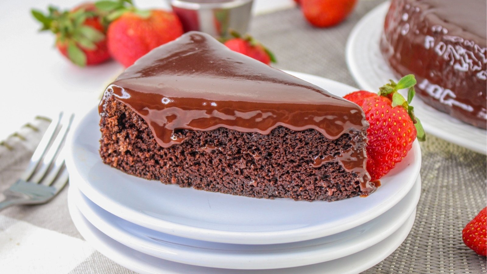 A slice of chocolate cake with glossy ganache sits on stacked white plates, surrounded by fresh strawberries.