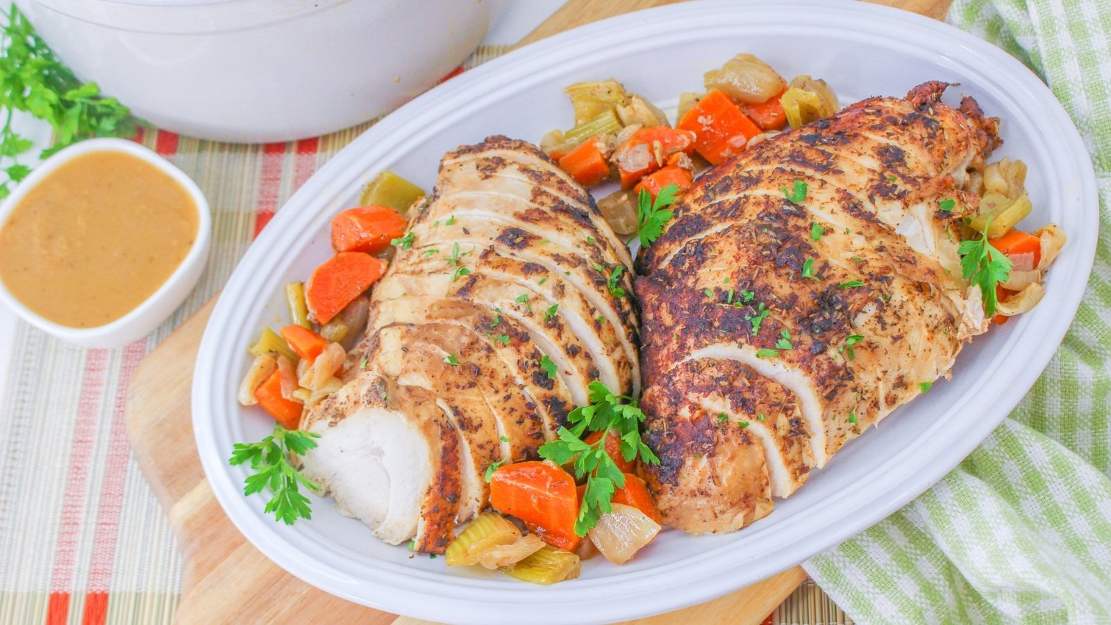 Sliced roasted chicken breast with herbs on an oval platter, surrounded by cooked carrots and celery. A small bowl of sauce is placed nearby.