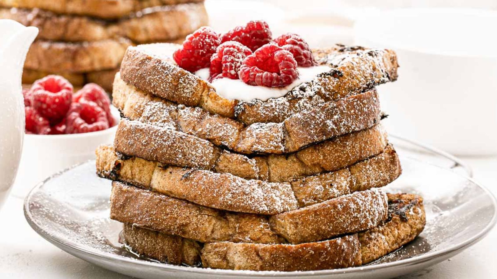 A stack of French toast topped with powdered sugar, creamy sauce, and fresh raspberries on a plate.