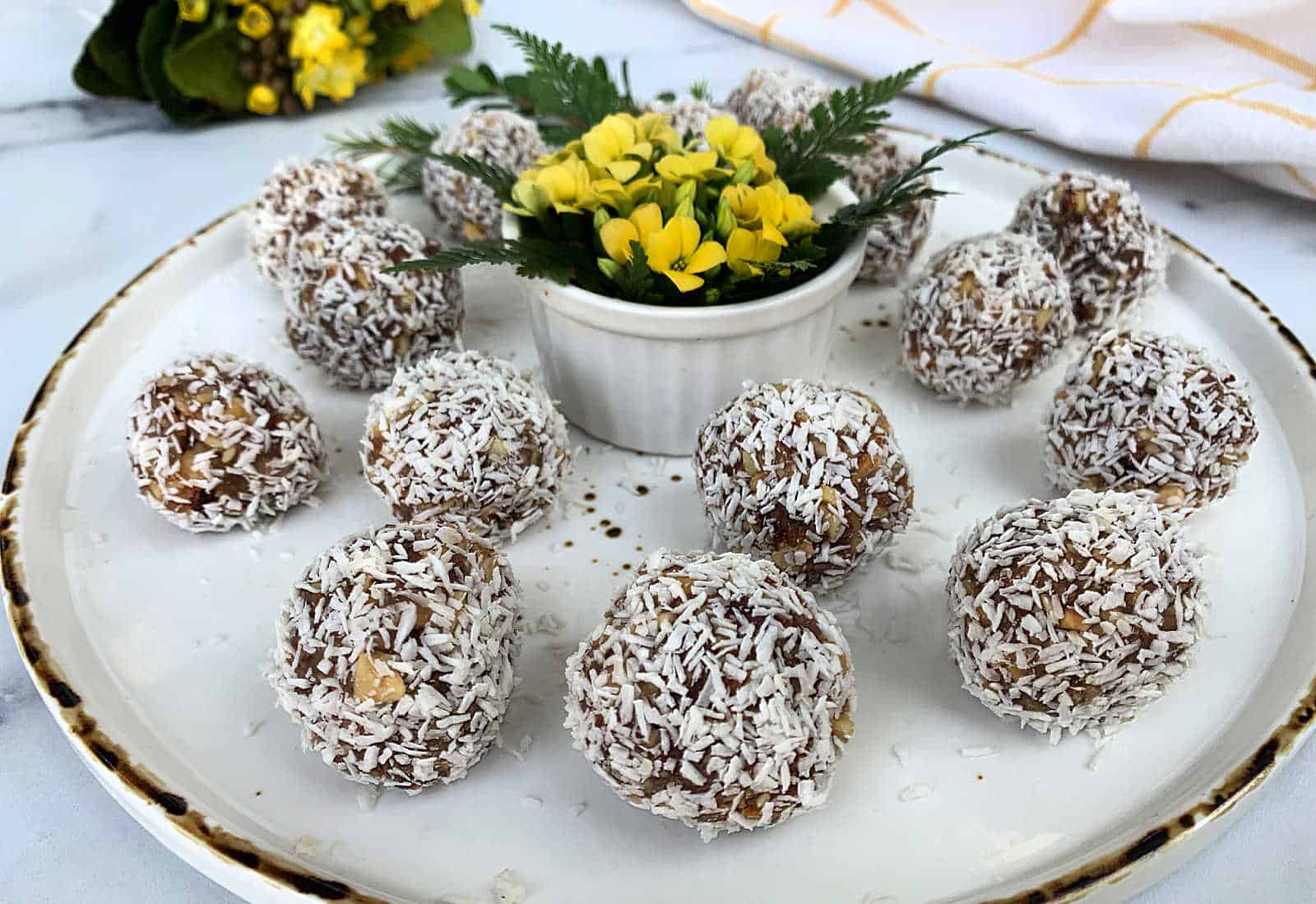 A plate of coconut-covered dessert balls arranged around a small dish with yellow flowers and green leaves.
