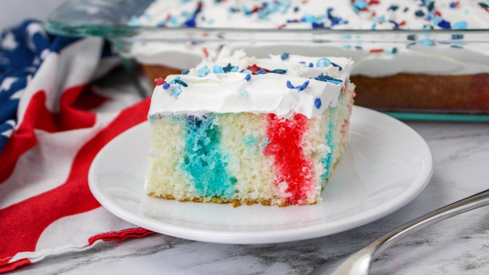 A slice of red, white, and blue poke cake topped with whipped cream and star-shaped sprinkles on a white plate, with a flag-themed cloth and more cake in the background.