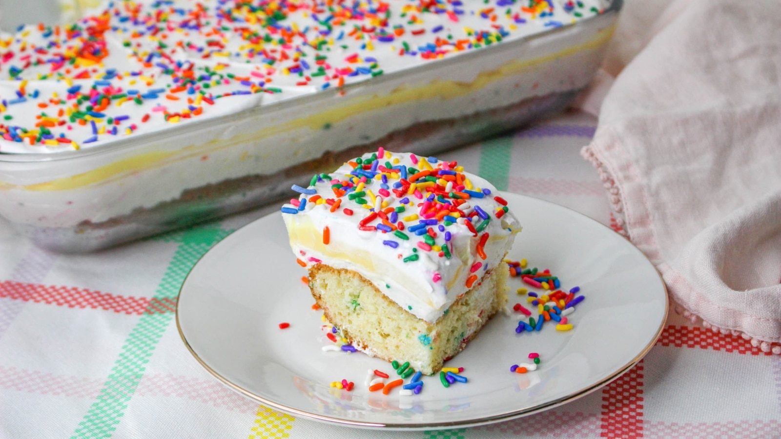 A slice of rainbow sprinkle cake with white frosting and additional sprinkles, served on a plate. A larger dish of the same cake is visible in the background on a checkered tablecloth.