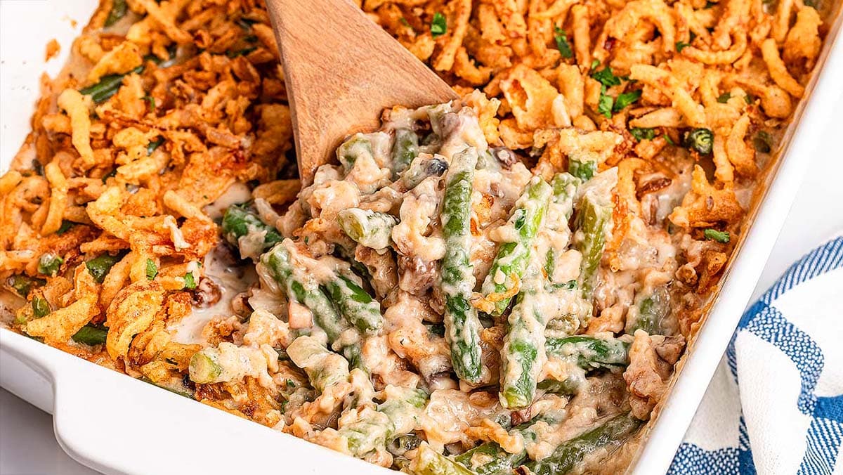 Green bean casserole in a white baking dish, topped with crispy fried onions, with a wooden spoon in the dish.