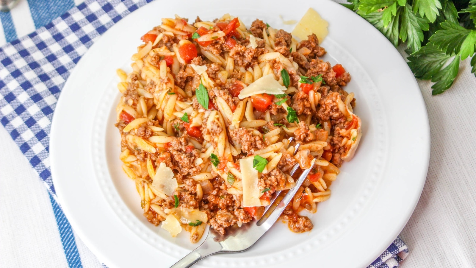A plate of pasta with minced meat, cherry tomatoes, and cheese shavings on a white plate. A fork rests on the plate, and a blue checkered napkin is underneath. Parsley is visible beside the plate.