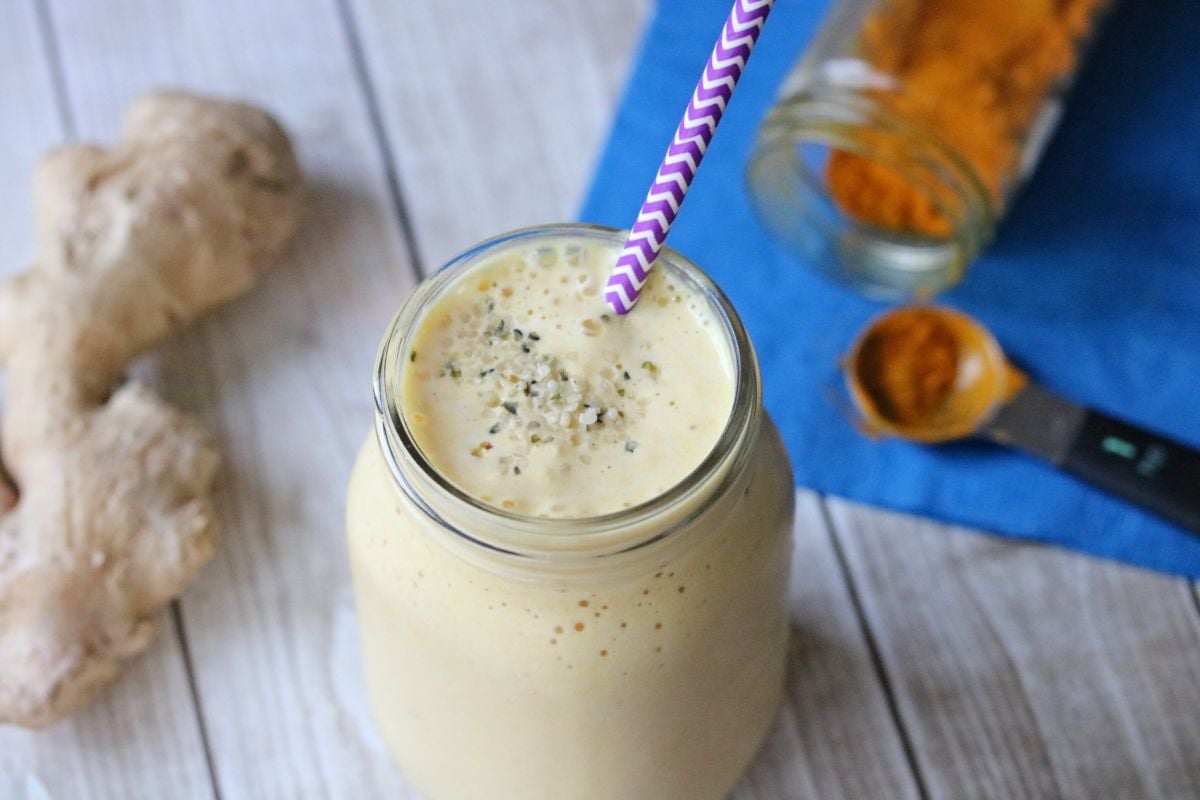 Turmeric peach smoothie in a mason jar with a purple straw. Turmeric root and powder and ginger root around it.