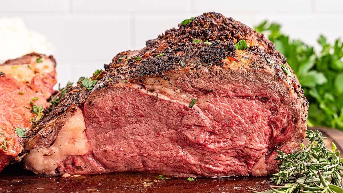 Close-up of a seasoned, cooked roast beef on a wooden board, garnished with fresh herbs. A sprig of rosemary and green leafy herbs are visible in the background.
