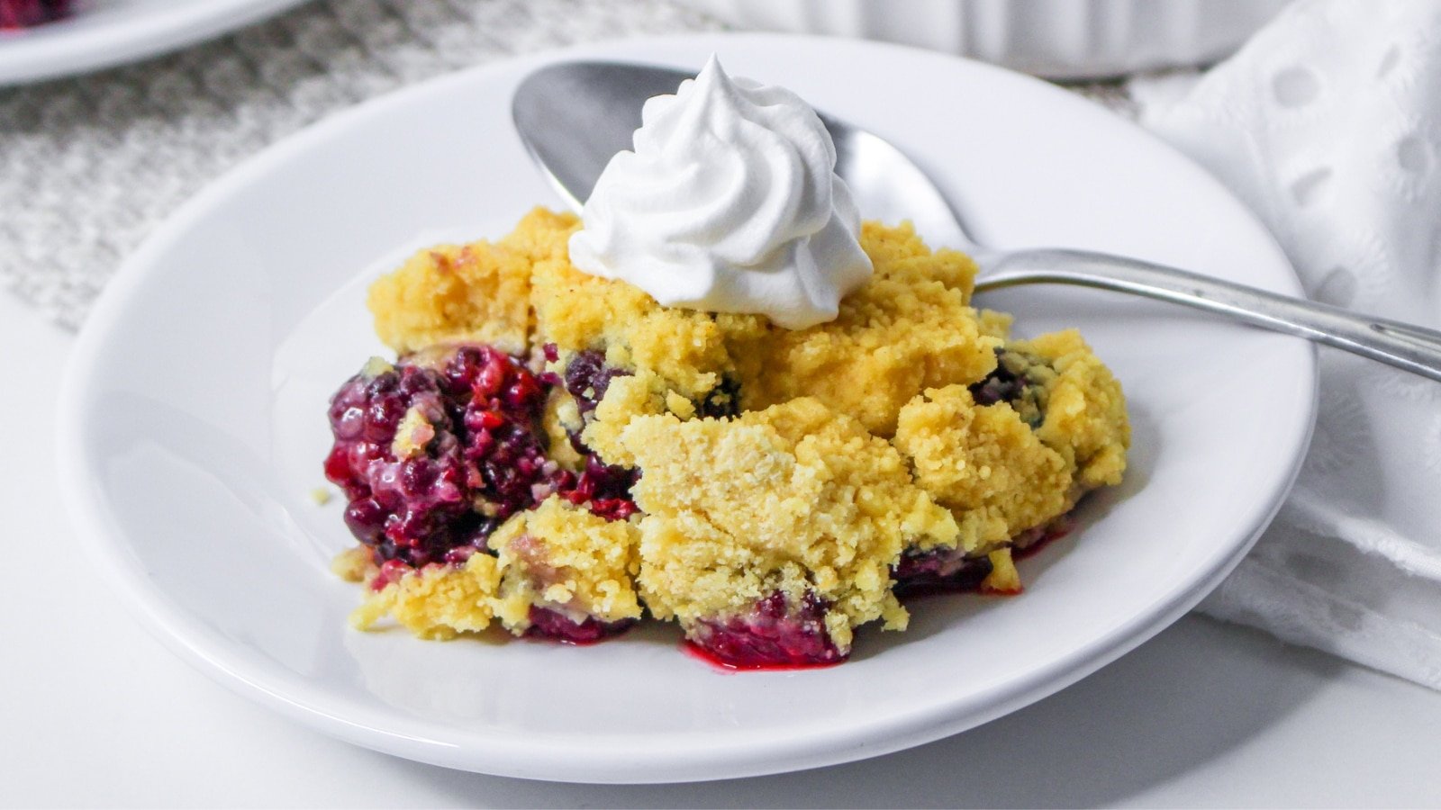 A dessert plate with a serving of berry crumble topped with whipped cream. A spoon rests beside it on the white plate.