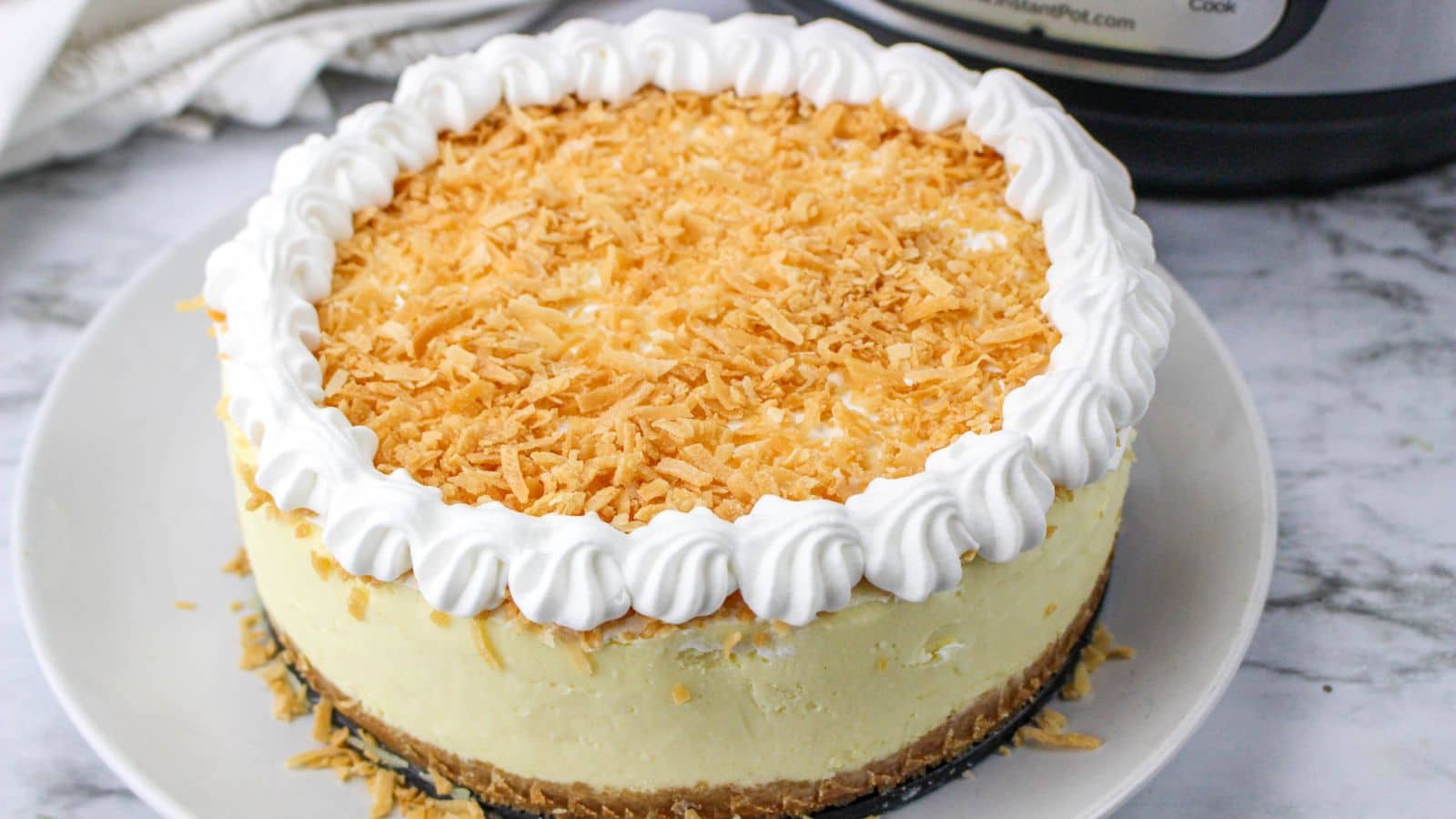 Cheesecake topped with whipped cream and toasted coconut on a white plate, set on a marble surface.