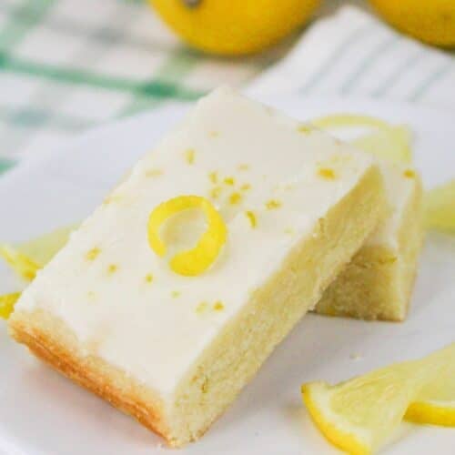 A slice of lemon cake with cream frosting, topped with a lemon twist, on a white plate. Lemon slices are nearby.