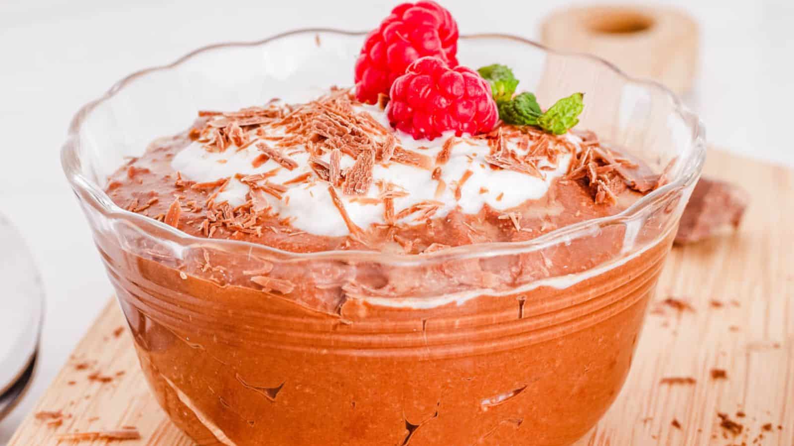 A bowl of chocolate mousse topped with whipped cream, chocolate shavings, two raspberries, and a sprig of mint.