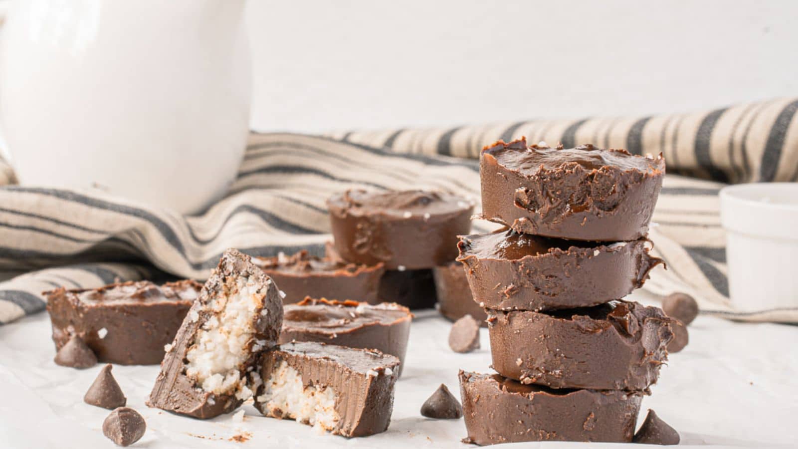 A stack of chocolate coconut fudge pieces is in the foreground. Some pieces are cut open, and a striped cloth and white jug are in the background.