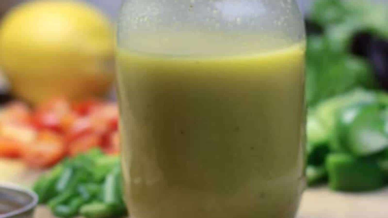 A jar filled with a yellow-green liquid, surrounded by assorted vegetables and a lemon in the background.
