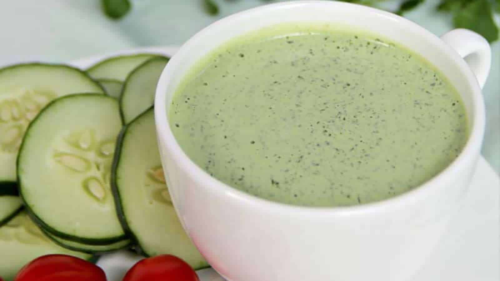 A white bowl filled with creamy green cucumber soup, garnished with cucumber slices and cherry tomatoes on the side.