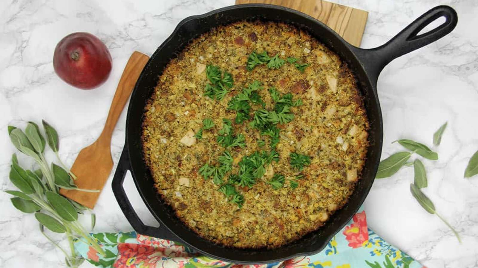 A skillet of baked stuffing topped with parsley, placed on a white marble surface. Nearby are a red apple, fresh sage leaves, a wooden spatula, and a floral-patterned cloth.